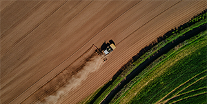 Aerial view of a tractor plowing a large field, creating parallel lines in the soil, with a strip of green vegetation bordering the field.